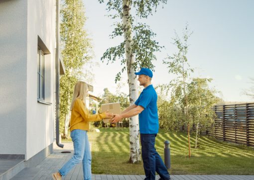 Delivery Man Delivers Cardboard Box Package to a Beautiful Young Woman, Who will Sign Electronic Signature POD Device. In the Background Cute Suburban Neighbourhood. Side View Shot