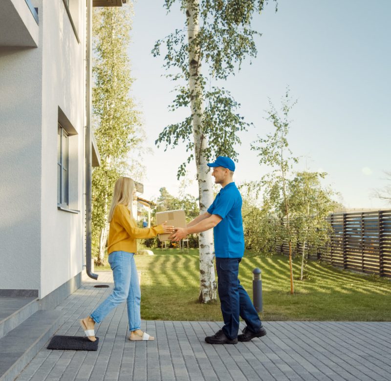 Delivery Man Delivers Cardboard Box Package to a Beautiful Young Woman, Who will Sign Electronic Signature POD Device. In the Background Cute Suburban Neighbourhood. Side View Shot