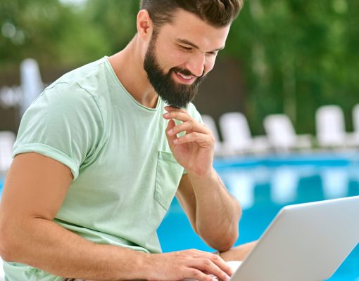 Working remotely. A young dark-haired man working on a laptop while sitting near the swimming pool