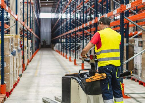 Warehouse worker wearing high visibility vest driving pallet truck between shelves in large distribution center