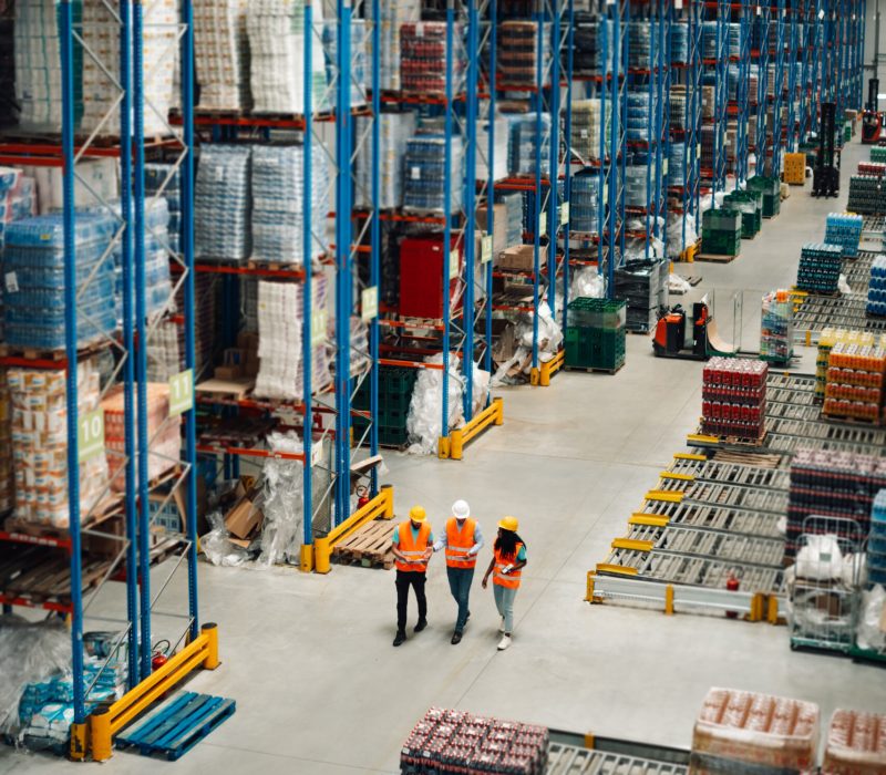 Three warehouse workers wearing safety vests and helmets are walking through large distribution center and controlling inventory using digital tablets